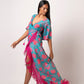 Walking pose of model in asymmetric blue floral wrap dress, one leg forward, showing fringe and flare sleeves in motion.