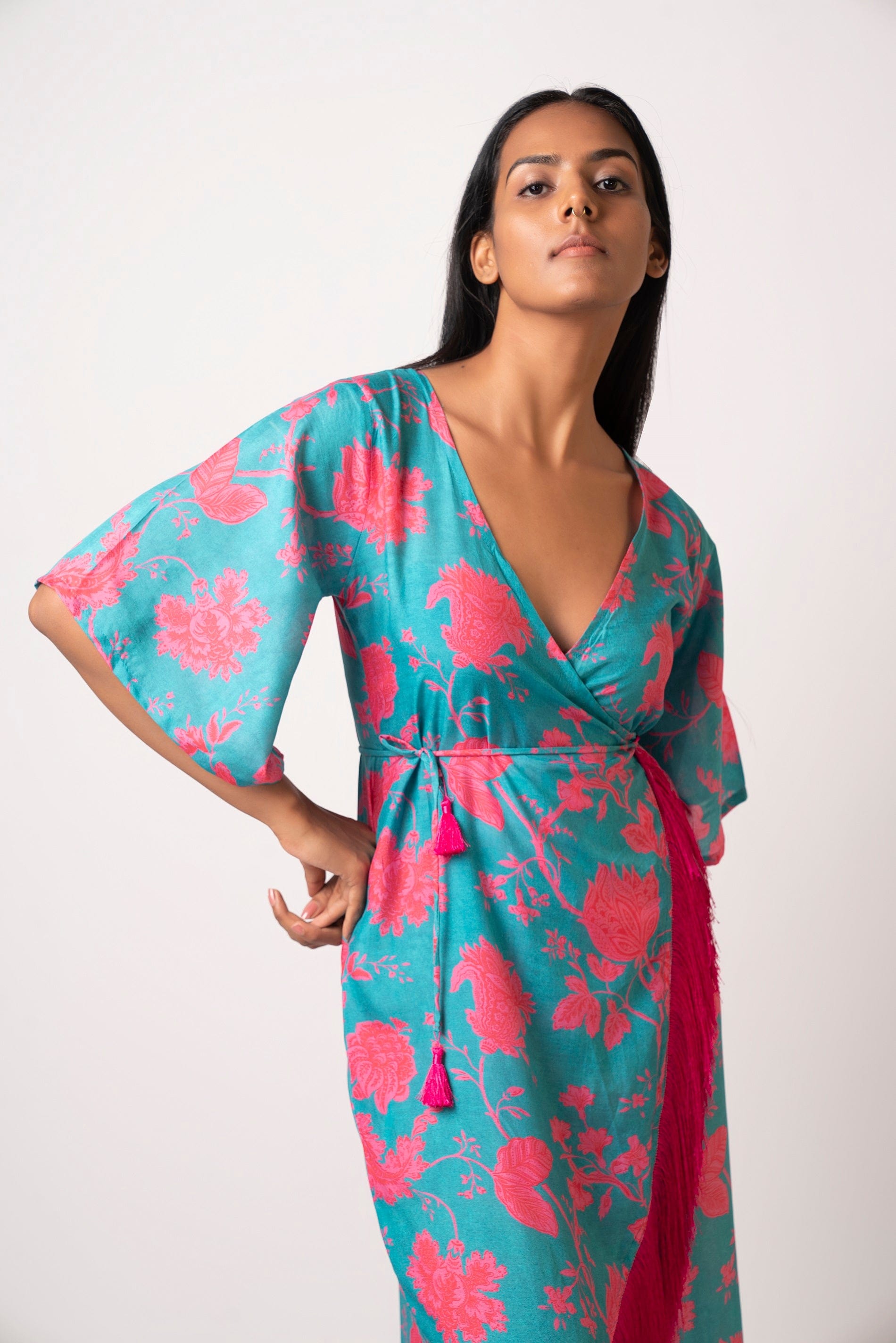Front-facing pose of model in floral blue muslin dress with side tie-up and flared hem, holding one hand at the waist.