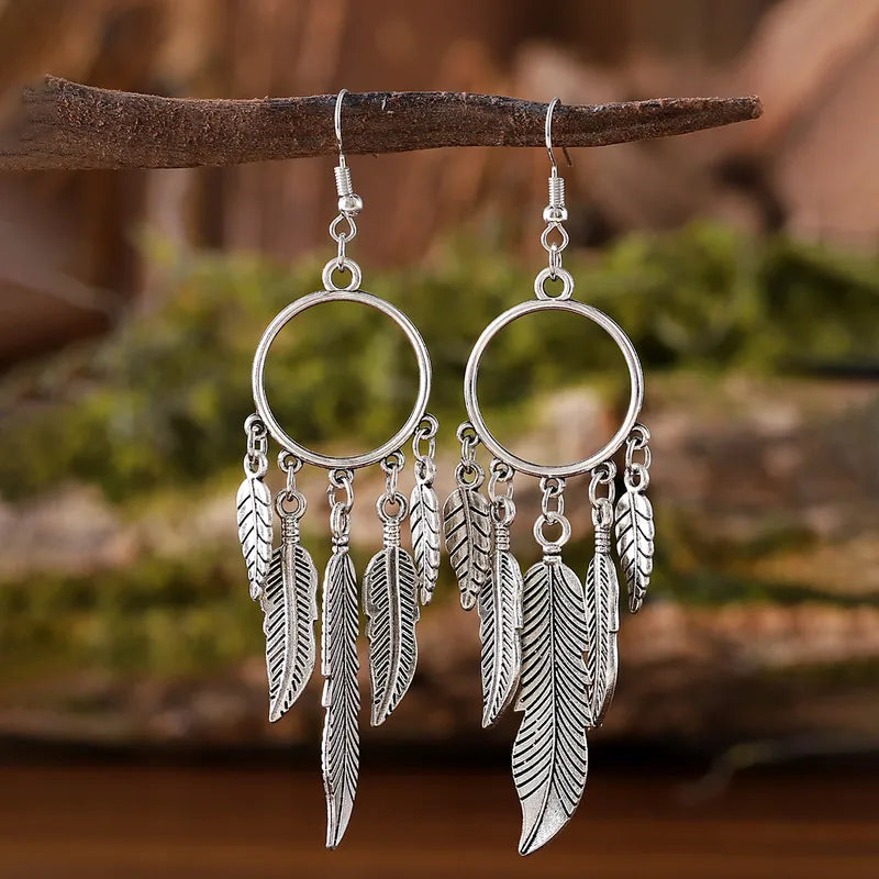A pair of antique silver Feather Hoop Drop Earrings with intricate metallic feather charms hanging from a circular hoop, displayed on a rustic branch background.