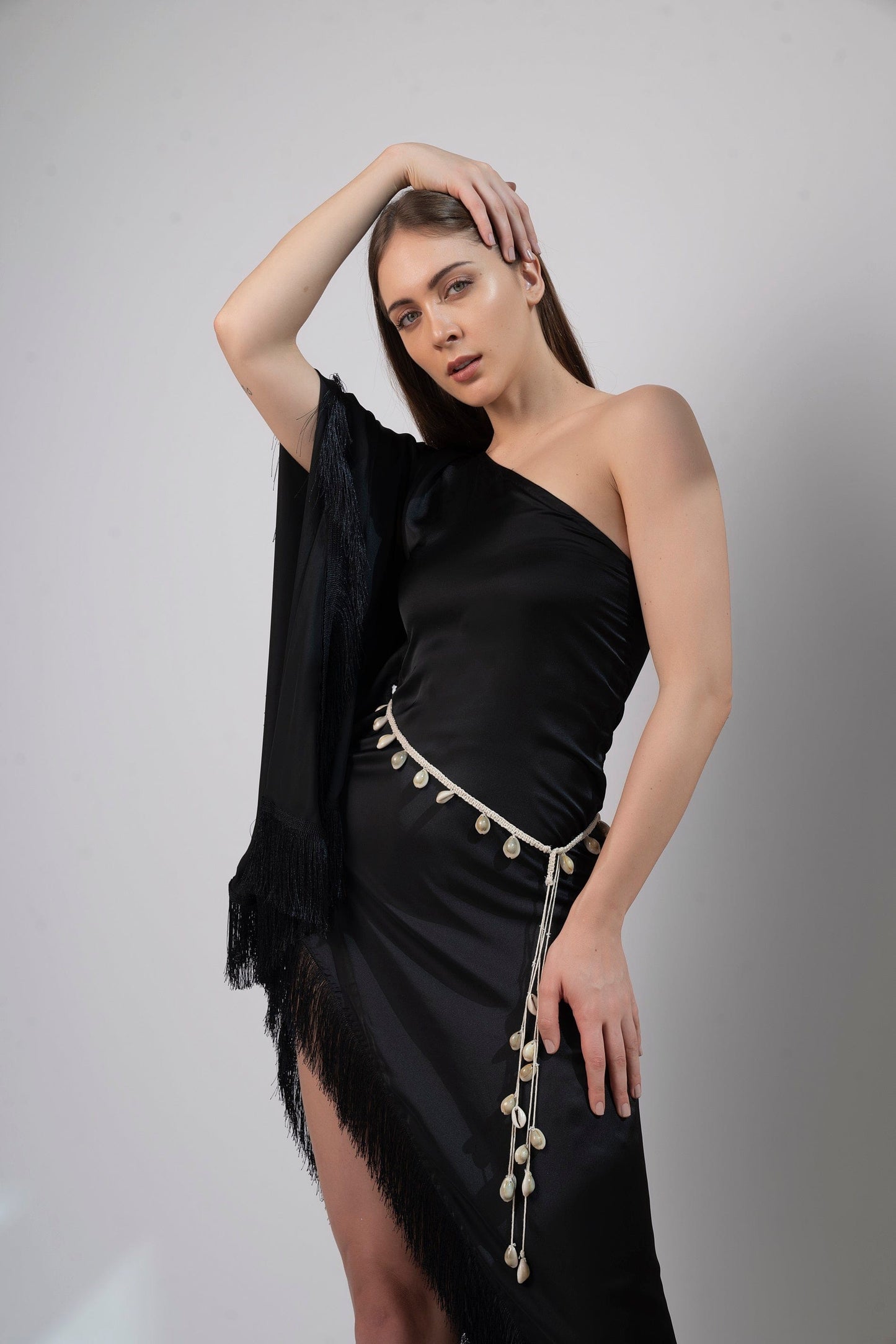 Zoomed-in slightly titled pose of black one-shoulder beach dress with fringes and tied waist belt.