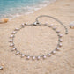 A Dainty imitation silver chain anklet with dangling white pearls laying on beach sand, perfect for summer vacations.